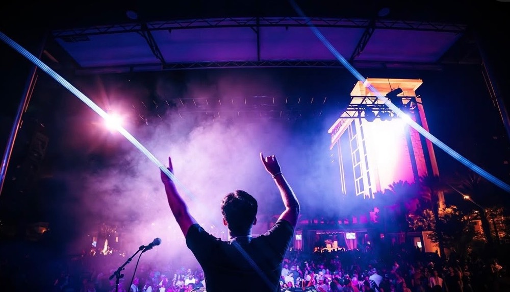 Performer with arms raised in front of a crowd under purple light and lasers